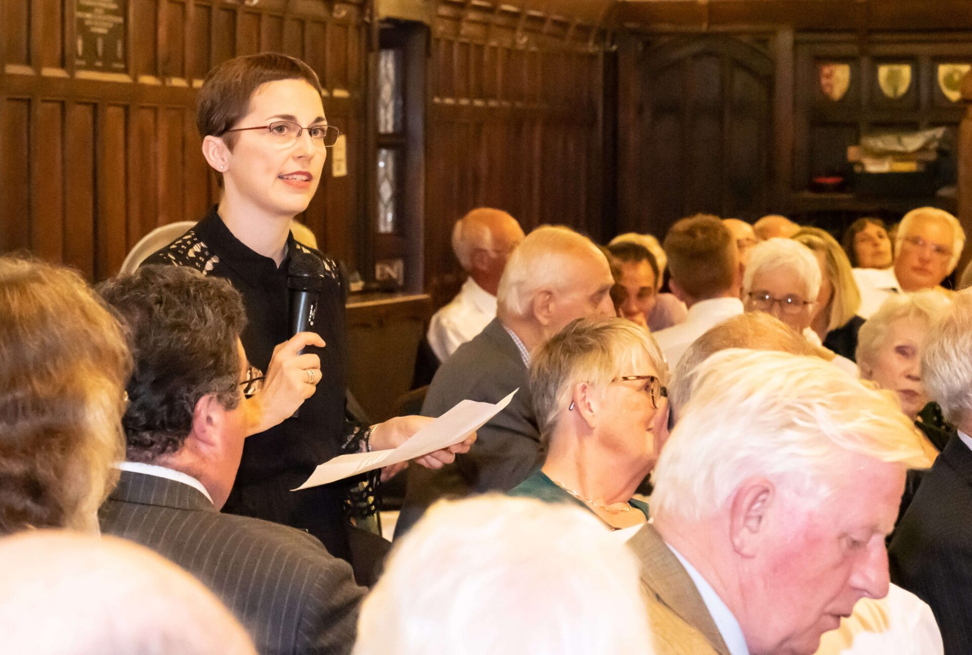 Annette Rubery stands with a microphone in a panelled room speaking to an audience.