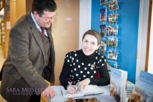 Annette Rubery is shown seated at a desk signing a book for a man who stands leaning on the table.
